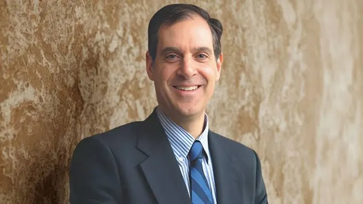 Fred Nathan Jr. smiles for a portrait wearing a dark suit, striped shirt and blue tie against a textured tan wall.