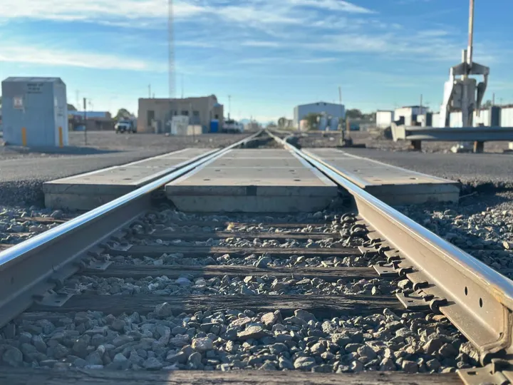 A ground-level view along railroad tracks, with gravel, concrete crossings and industrial buildings visible in the distance.
