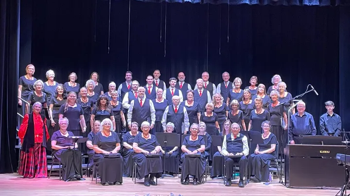 Members of the Mesilla Valley Chorale pose in rows onstage beneath a red curtain, wearing coordinated black outfits with red accents.