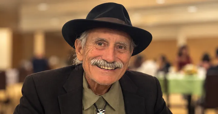 Max Adams, 78, smiles while wearing a black wide-brimmed hat, dark jacket, olive-green shirt and bolo tie against a softly blurred background.