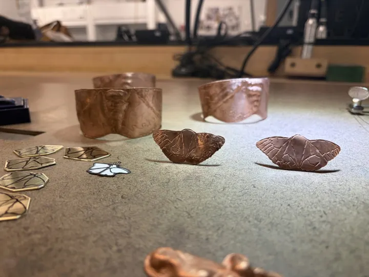 Handcrafted copper moth jewelry and textured metal cuffs by Piandria Shiner displayed on a worktable for NMSU's annual jewelry sale.