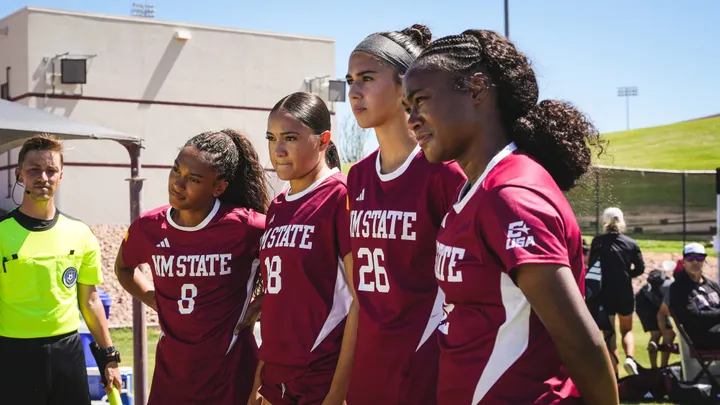 Four NMSU women's soccer players in crimson uniforms stand together on a sunny soccer field, with a referee at left and spectators in the background.