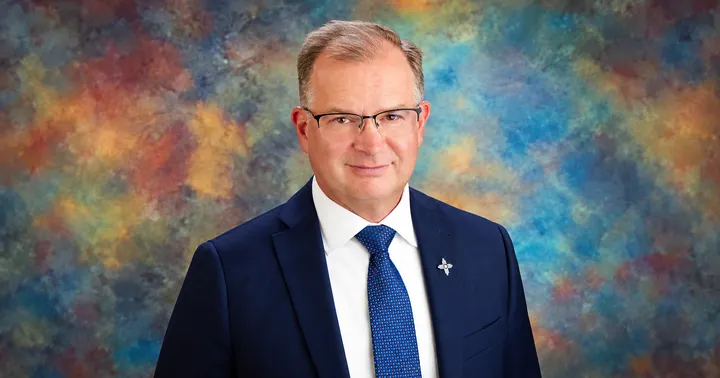 James Ellison, Republican candidate for New Mexico state treasurer, wearing glasses, a navy suit and blue patterned tie against a multicolored studio backdrop.