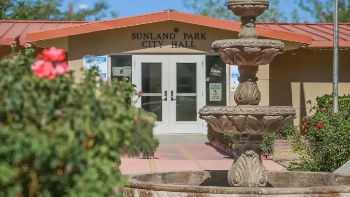Sunland Park City Hall's tan stucco exterior and red metal roof, with a decorative stone fountain, flowering bushes and glass entrance doors.