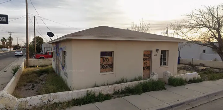 Boarded-up single-story building in Las Cruces with no-trespassing signs, graffiti and overgrown vegetation along a sidewalk.