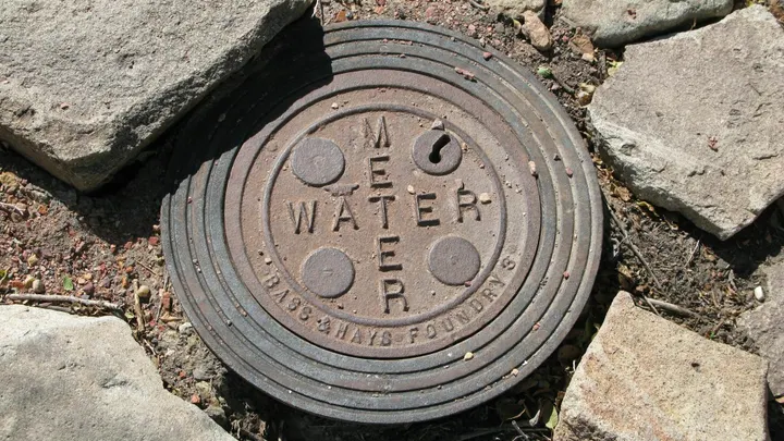 A round, weathered metal water meter cover with raised lettering sits in gravel, surrounded by large paving stones.