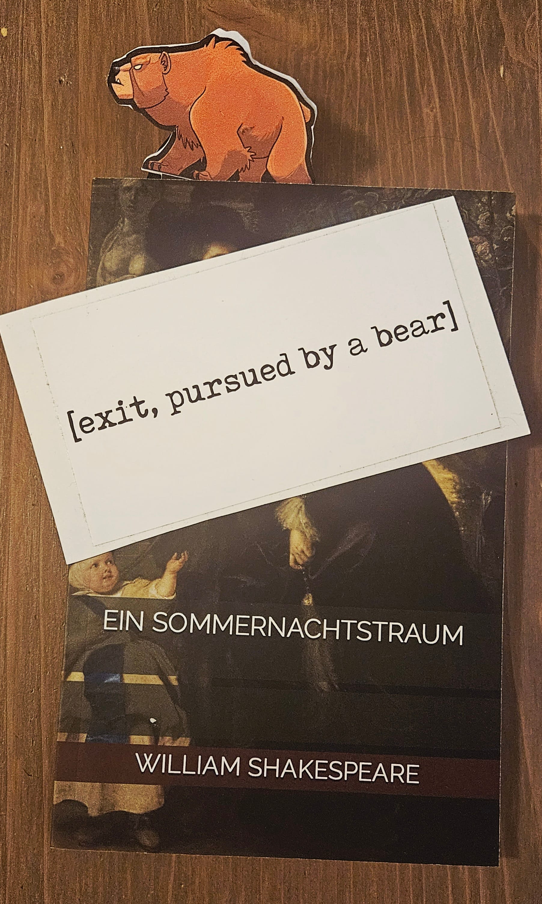 A paper figurine of a brown, grumpy grizzly bear stuck as a bookmark in a copy of Shakespeare's A Midsummer Night's Dream in German. A black-text-on-white unstuck sticker lies on top: [exit, pursued by a bear].