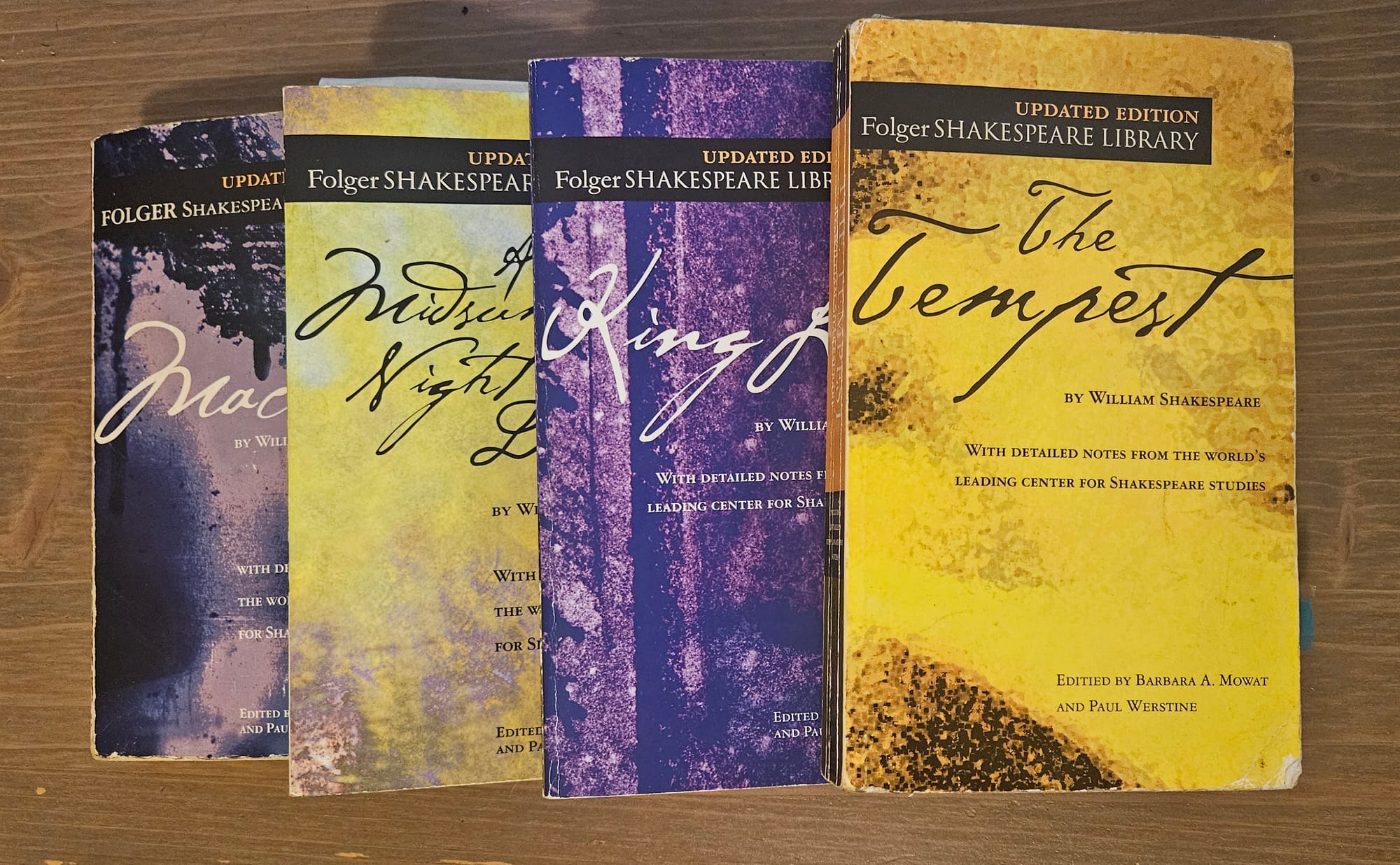 Four Folger editions of Shakespeare plays stacked slightly on each other, left to right: Macbeth (purple misty cover), A Midsummer Night's Dream (yellow-green), King Lear (purple stone), The Tempest (yellow).