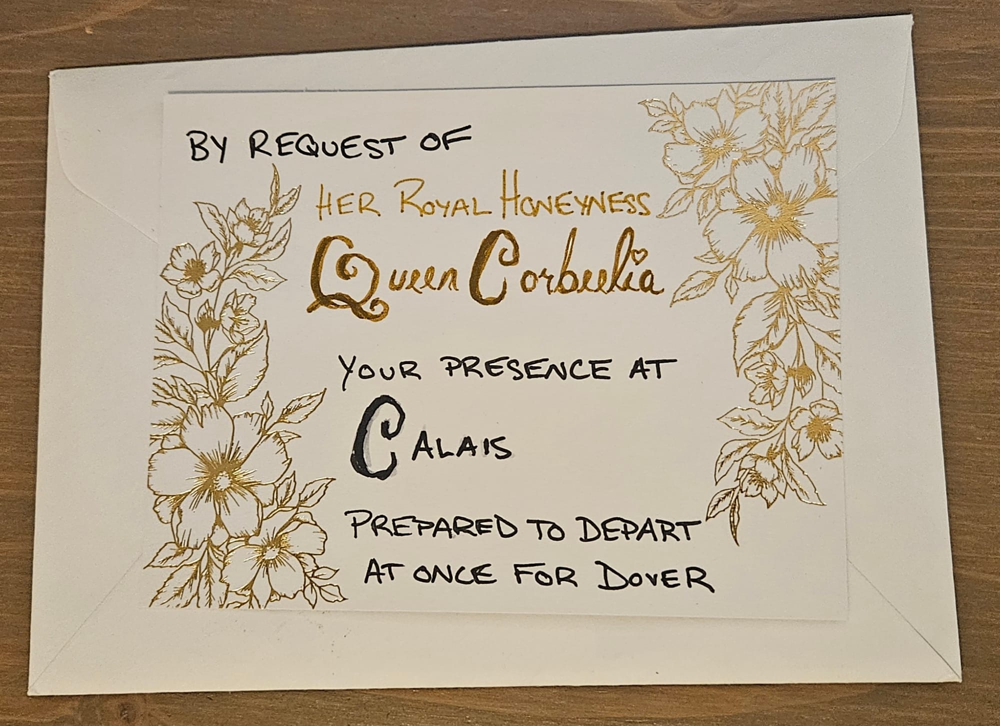 A white & gold wedding invitation that says, "by request of Her Royal Highness Queen Corbeelia, your presence at Calais, prepared to depart at once for Dover".