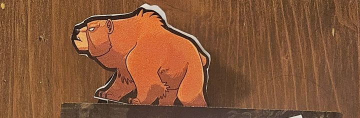 A paper figuring of a brown, grumpy grizzly bear, stuck as a bookmark in the top of a for-now unknown book.