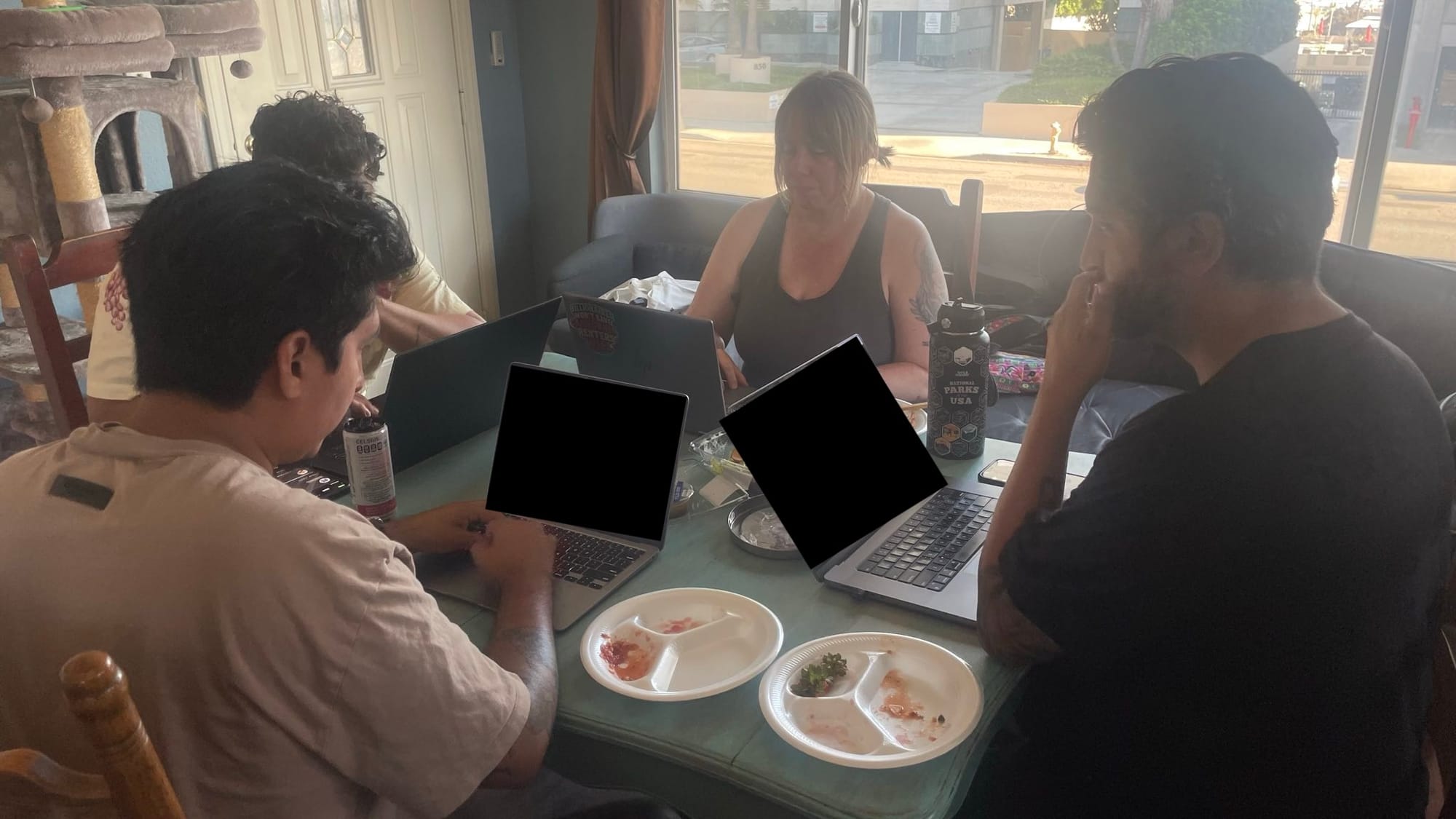 Four people sit around a table with laptops in front of them. Two of the visible laptop screens are redacted with a black square.