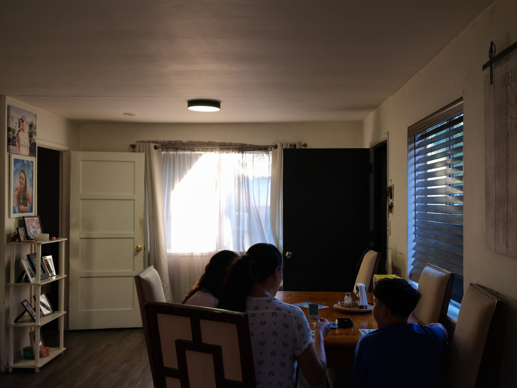 Inside of the family home, a mother and her two children play loteria on a dining room table. Their backs are to the camera and they are in the foreground.