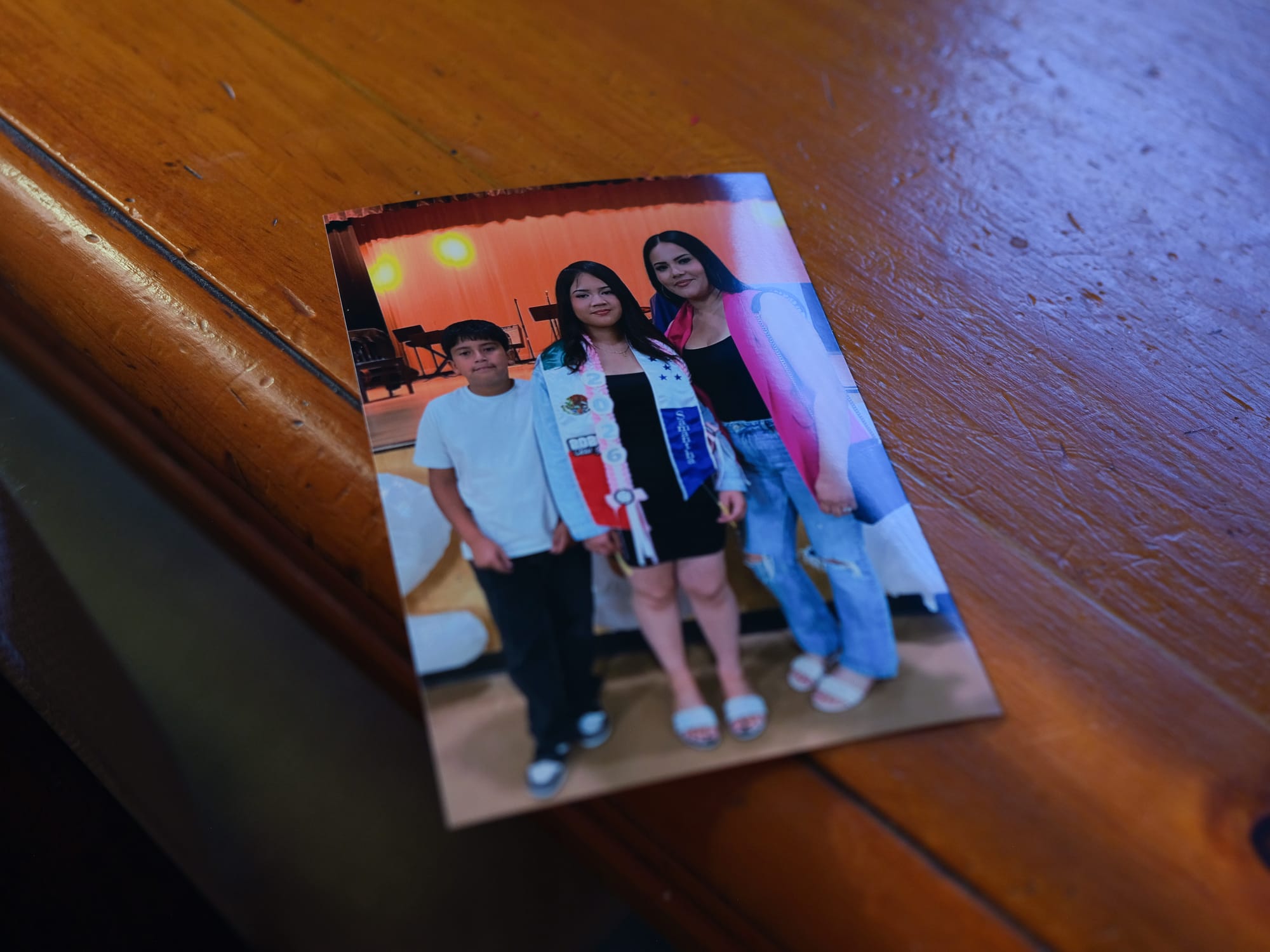 A young graduate is flanked by her younger brother and mother inside a school auditorium.