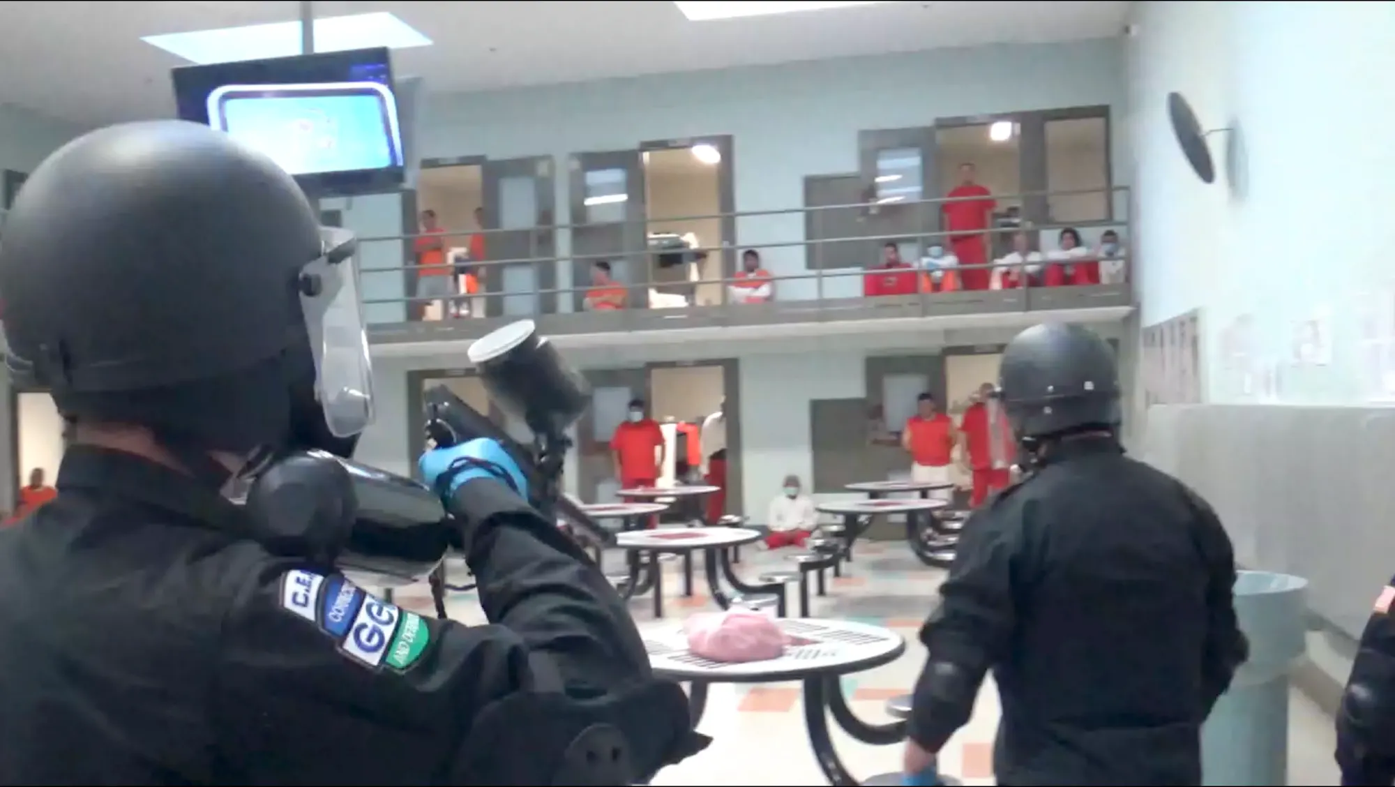 Several people in the background wearing red clothing, some sitting and some standing, as detention facility guard in the foreground points weapon at them. 