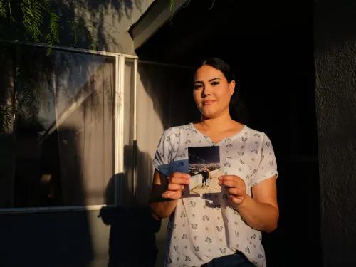 Woman with dark brown hair pulled back wearing white short sleeved shirt with repetitive blue and yellow pattern holds photo up in front of her chest. The photo is of her and her husband..