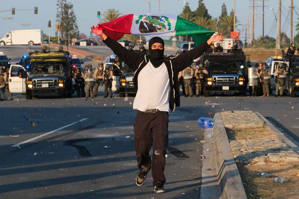 A person wearing a black mask holds a Mexican flag that is waving behind them. In the background, police trucks and police form a skirmish line.