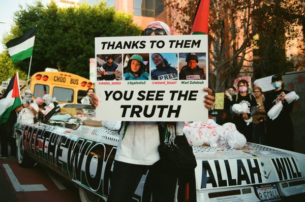 A protestor holding a sign in front of the bottom half of their face. The sign says "THANKS TO THEM YOU SEE THE TRUTH" and there are photos and names of Palestinian journalists.