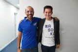 Two men standing indoors with their arms around each other's shoulders, smiling at the camera. The man on the left wears glasses and a blue polo shirt with Arabic script and "max" branding. 