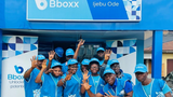 A group of Bboxx employees wearing blue caps and vests pose smiling in front of a blue storefront labeled "Bboxx Ijebu Ode", with several people holding up their hands to display fingers.