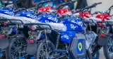 A fleet of new electric motorcycles parked indoors, primarily featuring blue models with the "Spiro" brand and "Ekon" model names printed on the fuel tank area, alongside some red motorcycles