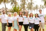 Rian and team members standing together outside on a lawn with palm trees and thatched-roof buildings in the background during their 2024 retreat, all wearing white branded T-shirts.