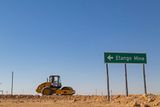 A yellow road roller operating on a dusty construction site near a green directional sign reading "Etango Mine" pointing left against a clear blue sky.