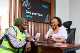 A Watu customer in a high-visibility safety vest and beanie smiling while consulting with a representative across a desk in an office with promotional branding.
