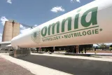 A large white industrial storage tank featuring the green "omnia" brand logo alongside the text "NUTRIOLOGY • NUTRILOGIE*", set against a blue sky with light clouds and industrial silos.