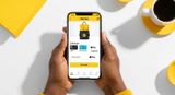 Hands holding a smartphone displaying the Nomba app checkout interface against a bright yellow and white background with a coffee cup.