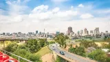 Panoramic view of the Johannesburg city skyline under a partly cloudy sky, featuring elevated highway bridges, green spaces, and vehicular traffic in the foreground.