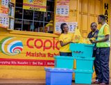 A female Copia agent in a yellow shirt standing at a brightly painted yellow storefront, receiving yellow shopping bags from a delivery worker.