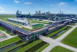 Aerial view of a sprawling industrial refinery complex featuring extensive blue and multi-colored pipe racks, processing units, and water channels under a blue sky.