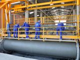 Industrial workers in blue uniforms and hard hats operating heavy machinery inside a manufacturing facility, with a large concrete pipe section in the foreground.