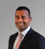 Professional headshot of Mesh Pillay, CEO of YW Capital, smiling in a dark suit, white collared shirt, and pinkish tie against a neutral grey background.