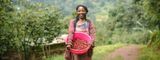 A smiling female smallholder coffee farmer holding a pink basin filled with harvested red coffee cherries outdoors on a rural mountain farm.