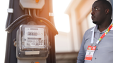 Stephen Adwong'a, CEO of Miale Solar, wearing a conference speaker badge, stands beside an installed analog watthour electricity meter.