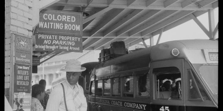 A black man waits for a bus under a sign that reads "Colored Waiting Room."