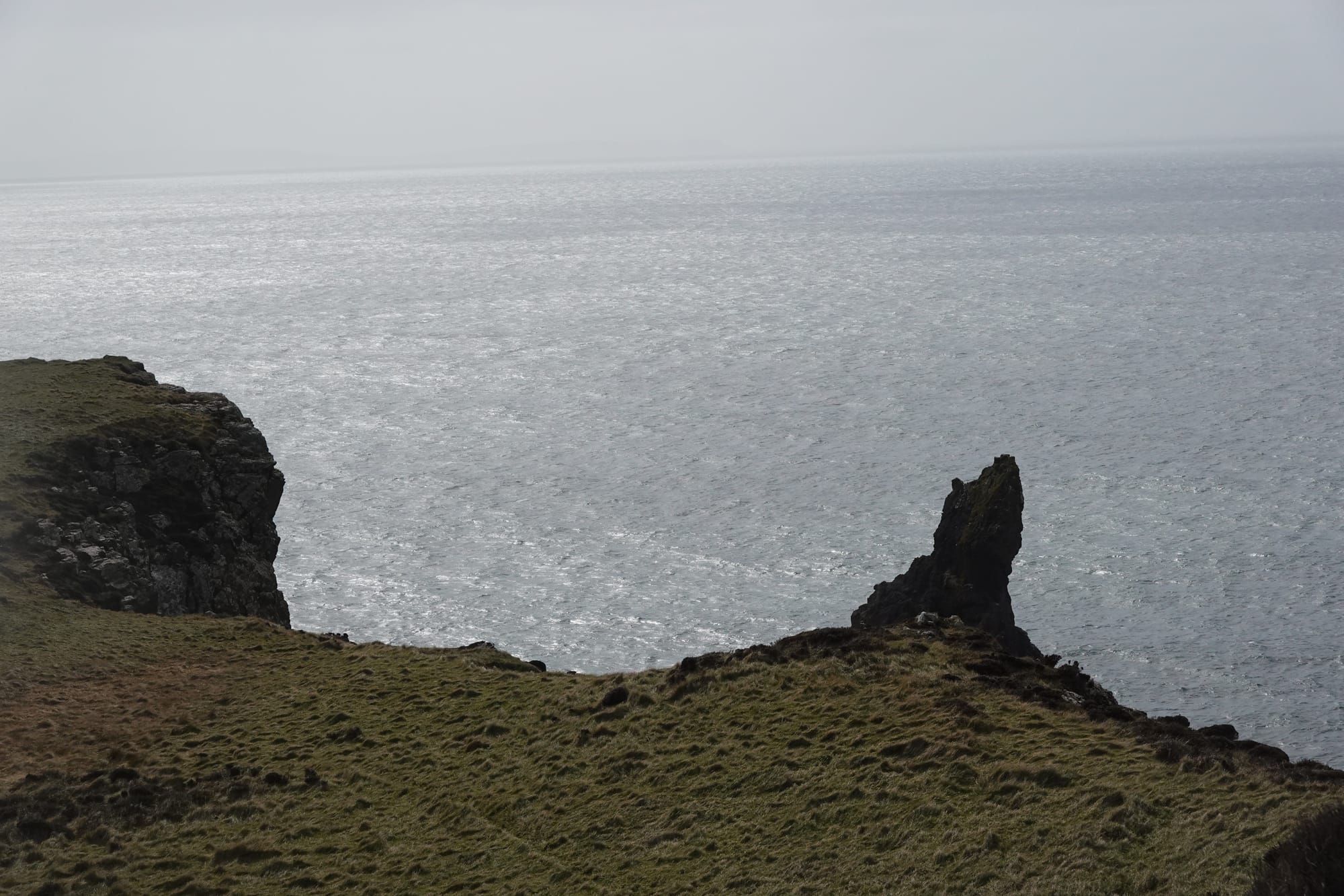 Grassy coastal cliff slopes down toward the sea, with a sharp rock formation jutting up near the edge.