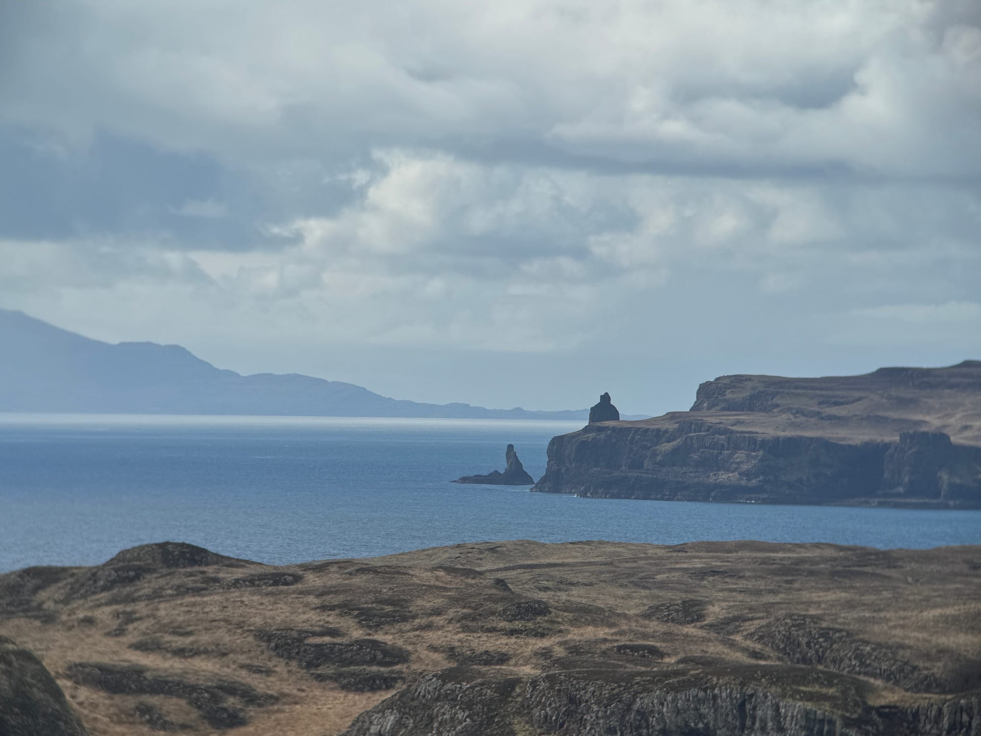 Wide coastal landscape with rugged cliffs and distant sea stacks beneath a cloudy sky, overlooking calm blue water.
