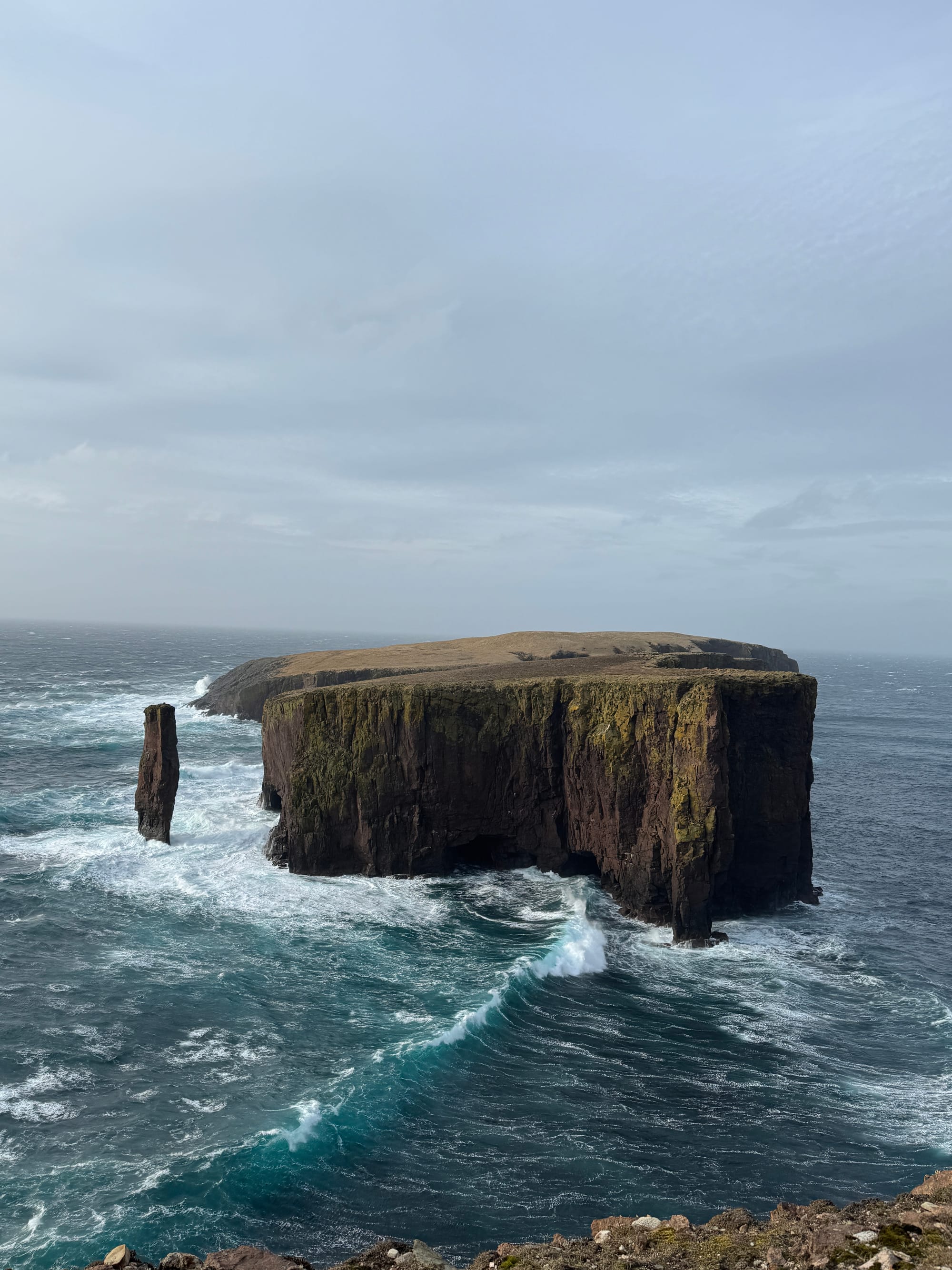 A rugged sea stack rises from churning blue-green water, with waves crashing against steep, rocky cliffs beneath a cloudy sky.