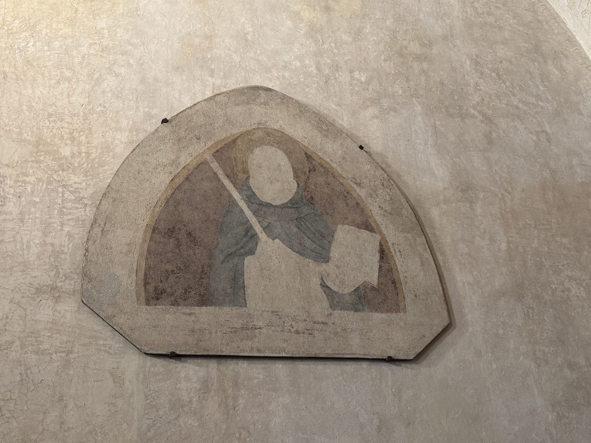 A close-up of a fresco painted directly on a plaster wall, depicting a figure within an octagonal frame. The figure is partially visible, holding a long object and a rectangular item, possibly a staff and a tablet. The colors are muted, primarily light gray and blue, with signs of age and wear on the wall, including cracks and areas of faded plaster.
