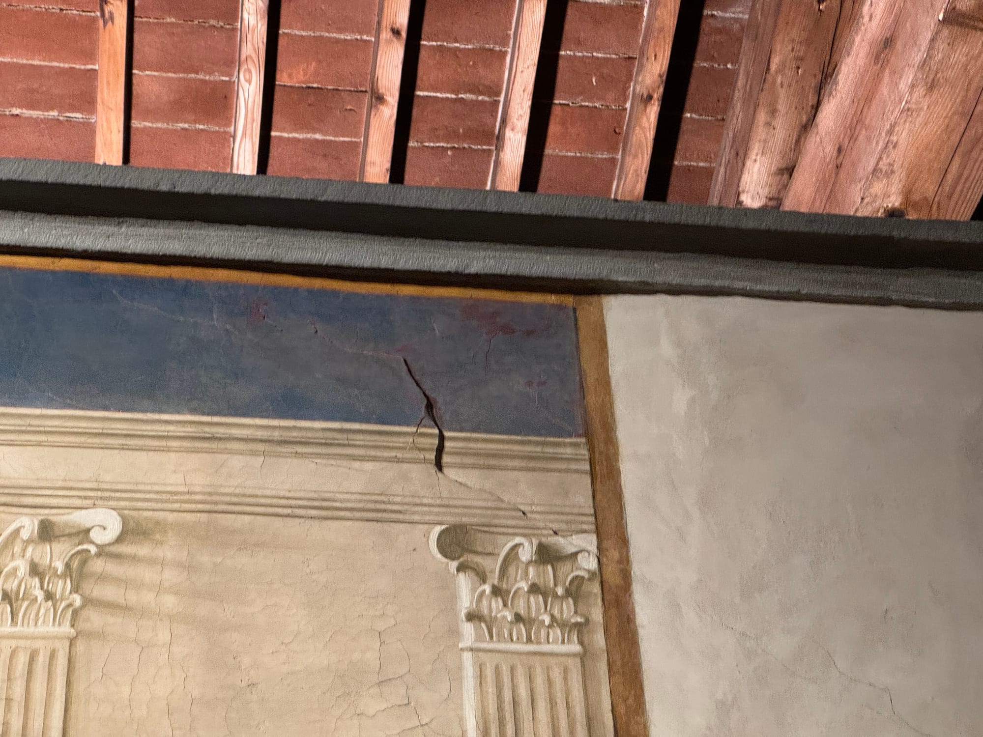 A section of a wall showing the upper portion of a fresco beneath an exposed wooden roof. The wall has a rectangular panel with a blue background and a partially visible architectural frame painted in light tones. The wooden beams of the ceiling are visible above, showing a warm reddish-brown color and a rough texture. Cracks are visible in the painted plaster along the edges.
