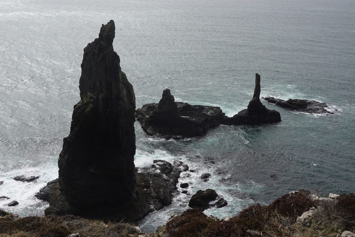 Tall, jagged sea stacks rise from rough ocean waters near a rocky shoreline under overcast light.