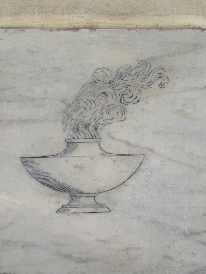 A close-up photograph of an engraving on a marble surface depicting a classical oil lamp. 
