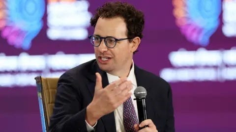Reuters Dario Amodei, Anthropic's CEO, speaking on stake while holding a microphone, wearing a black suit jacket, white shirt and red and navy spotted neck tie.