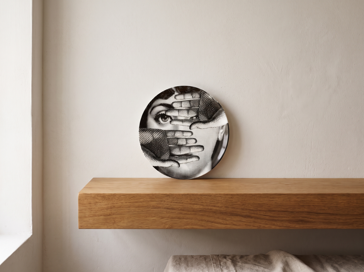 Fornasetti "Tema e Variazioni" porcelain plate with illustrated woman's face partly covered by hands, displayed upright on an oak shelf against a plaster wall.