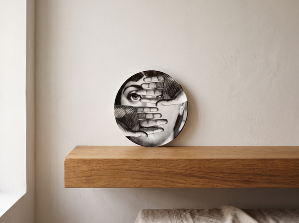 Fornasetti "Tema e Variazioni" porcelain plate with illustrated woman's face partly covered by hands, displayed upright on an oak shelf against a plaster wall.