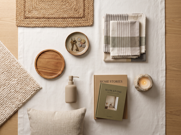Centered flat-lay of styled home essentials — tea towels, ceramic dish, trivet, cushion, candle and books — on warm white linen and oak, editorial interior photography.