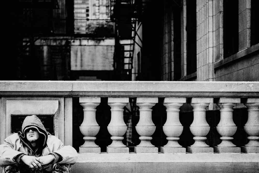 Homeless person in hooded jacket by ornate railing, a black and white photo
