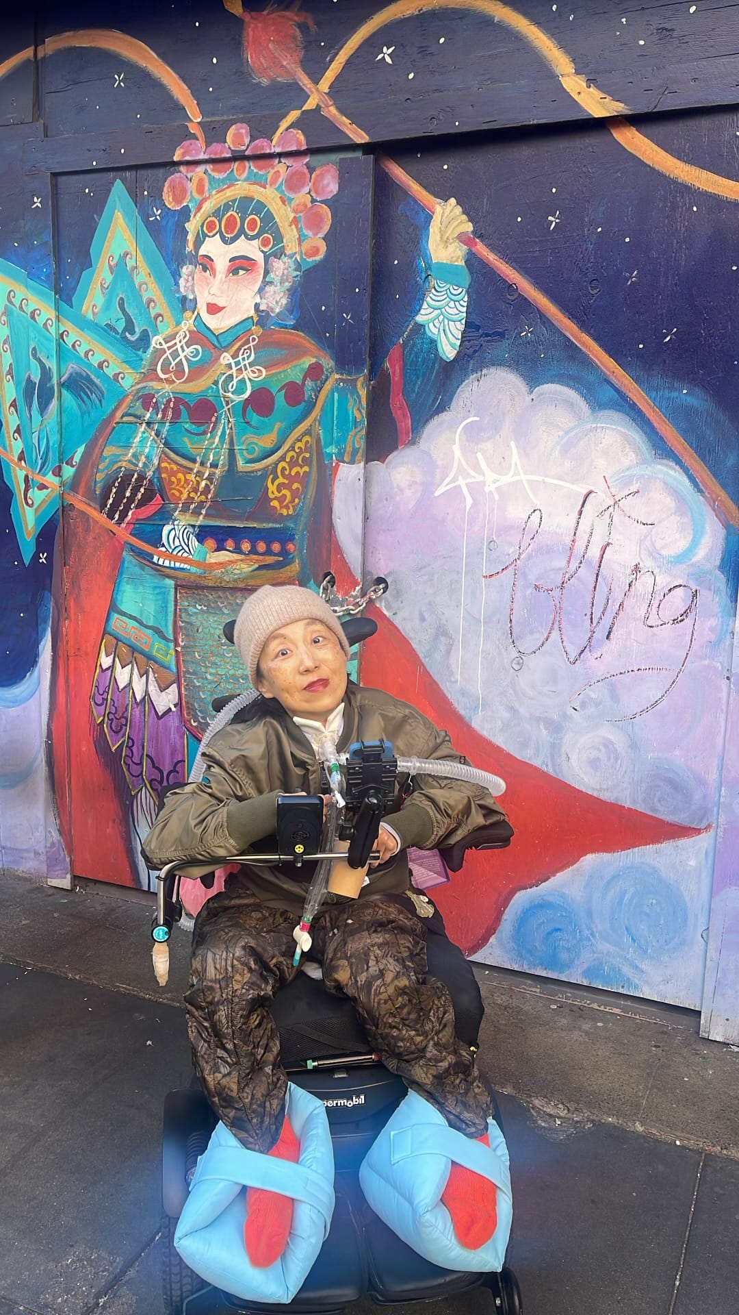 a photo of me, an Asian American woman in a wheelchair with a tracheostomy and a tube attached to a ventilator. I’m wearing a beanie, olive green jacket and camouflage pants. Behind me is a mural featuring a Chinese opera singer